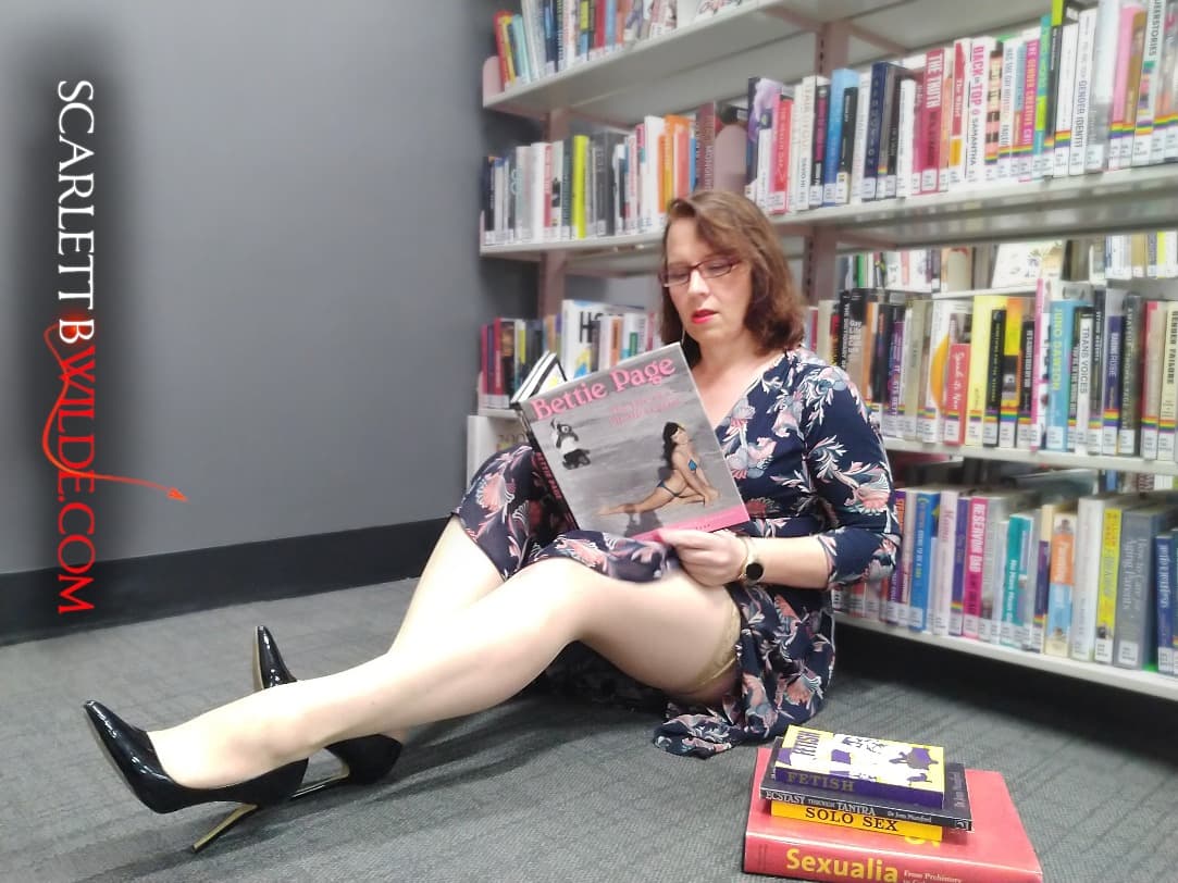 Scarlett B Wilde shows off fantastic legs in tan stockings and black heels while reading a Betty Page book. She's in the sexuality section in a library.