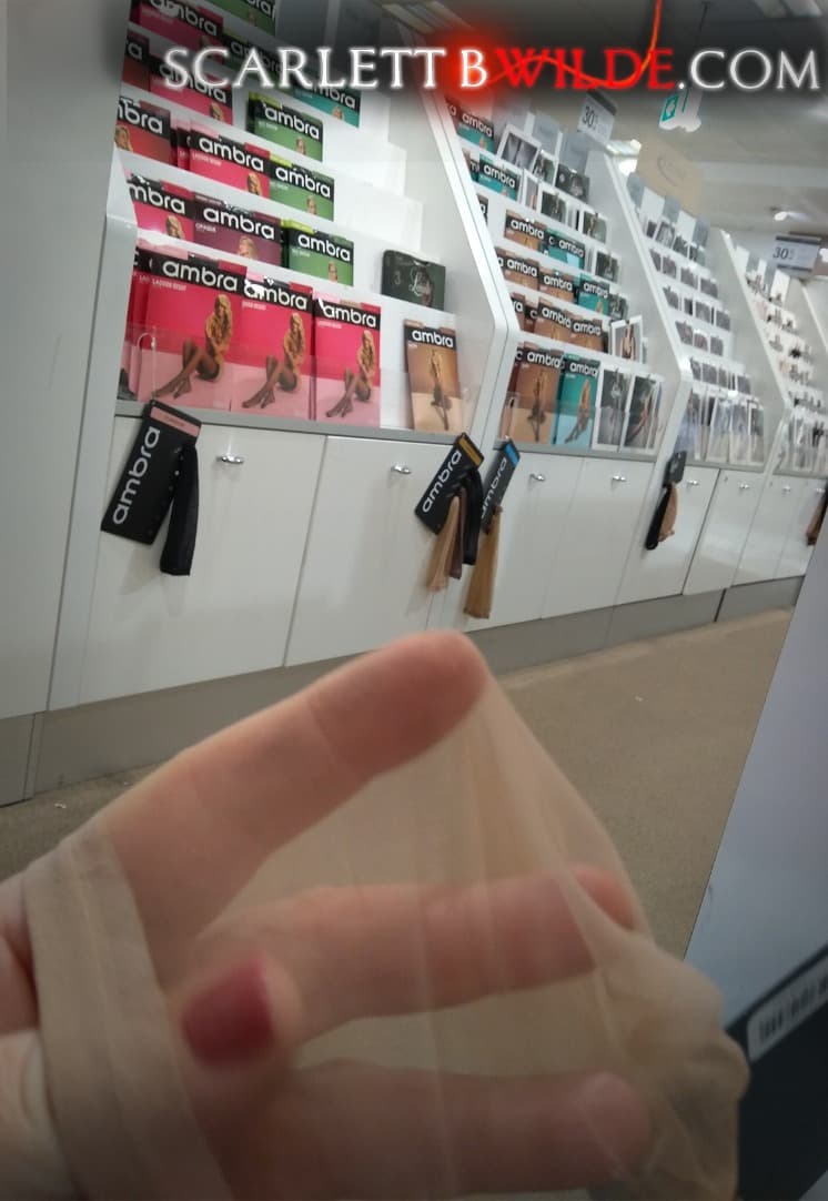 Scarlett B Wilde holds a sheer suntan pantyhose in between her fingers in the hosiery section of a retail store. Packets of pantyhose can be seen in the background.