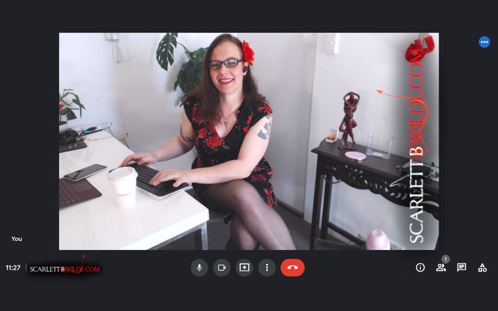 Scarlett B Wilde sits at a office desk looking into the webcam with a smile and wearing glasses. 
The short floral dress shows off her black stockinged legs.