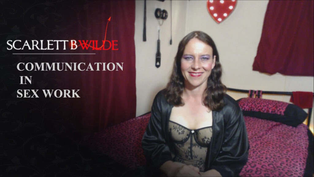 Video still of Scarlett B Wilde talking in a vlog about communicating with her clients. She sits on bed wearing Black teddy and satin black robe.