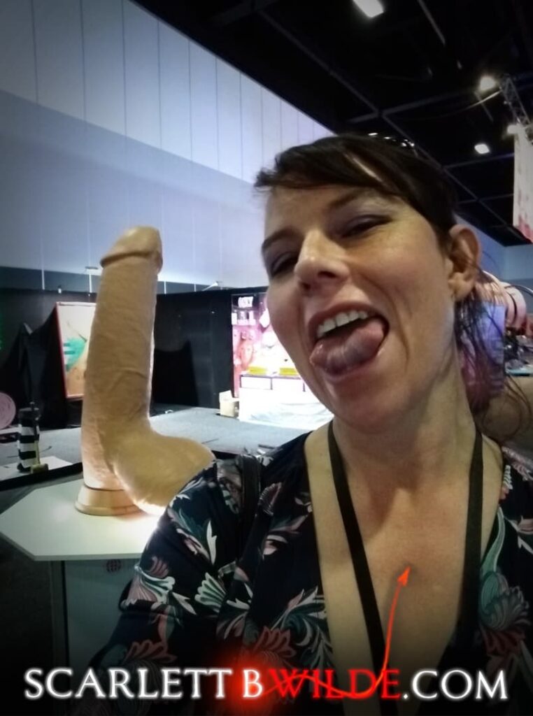 Scarlett B Wilde stands next to a giant dildo display at Sydney Sexpo 2018. She folds over her amazing tongue without much trouble